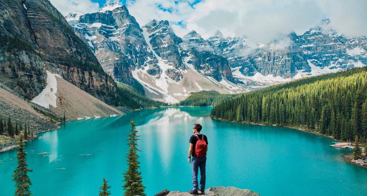 Una persona contemplando un lago turquesa rodeado de dramáticas montañas cubiertas de nieve.