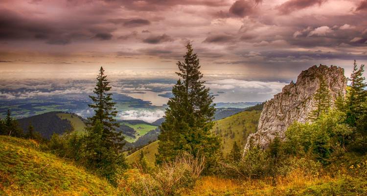 Dramatischer Bergrücken und Wald rahmen einen entfernten See unter düsteren, farbenfrohen Morgenrot-Wolken ein.