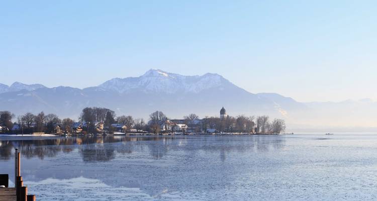Ruhiger See mit Spiegelung von Winterbäumen und verschneitem Bergpanorama im Hintergrund sowie einer kleinen Siedlung am Ufer.