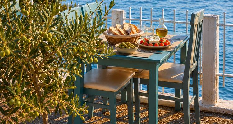 Table côtière confortable avec olives, pain, tomates et vin blanc disposés près d'un olivier donnant sur une eau bleue scintillante.