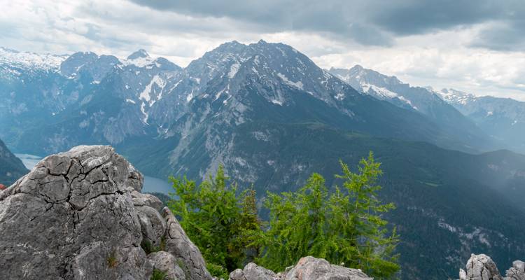 Dramatische Alpenpanorama mit zerklüfteten grauen Felsen im Vordergrund, immergrüne Bäume und aufragende schneegestreifte Gipfel unter einem stimmungsvollen Himmel zur Mittagszeit.