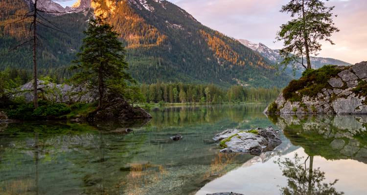 Ruhiger Alpensee mit kristallklaren Spiegelungen, immergrüne Bäume und sonnenbeschienene Berghänge während der goldenen Stunde.