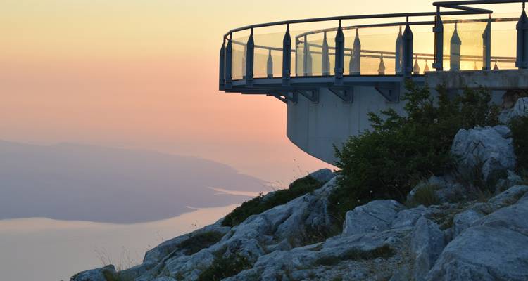 Mirador de cristal se extiende sobre una ladera montañosa al atardecer, resplandeciendo sobre la costa del Adriático.