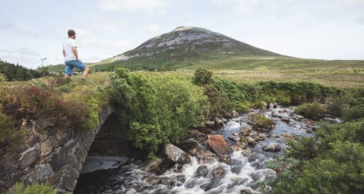 Une personne debout sur un pont près d'un ruisseau, avec une montagne en arrière-plan.