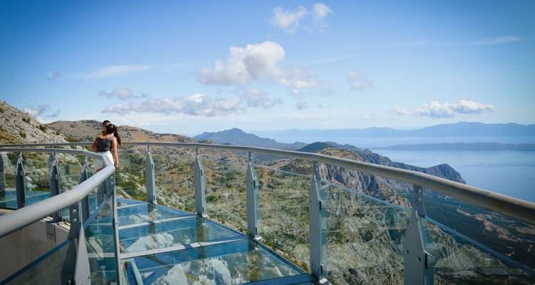 Una mujer se sienta en el Biokovo Skywalk de cristal con vista a las espectaculares montañas costeras y el Mar Adriático.