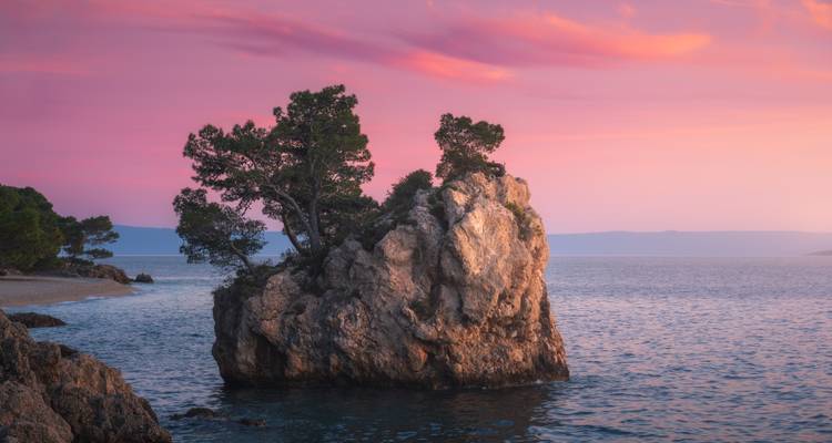 La icónica isla de piedra de Brela coronada de pinos brilla bajo un vibrante cielo de atardecer rosa.