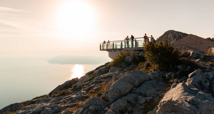 Bezoekers verzamelen zich op een glazen skywalk die uitsteekt boven een dramatische bergflank aan de kust tijdens de zonsondergang.