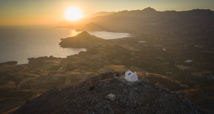 Le soleil se couchant sur la mer de Libye projette une lumière dorée sur la côte crétoise accidentée et une minuscule chapelle blanche perchée sur une crête de montagne.
