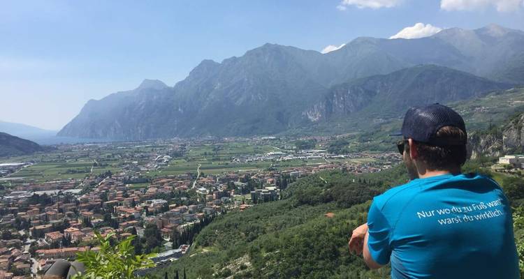 Un voyageur vêtu d'une chemise bleue observe une vaste vallée du lac de Garde encadrée par les montagnes.
