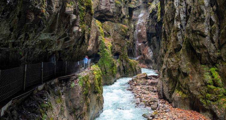 Gorge rocheuse étroite avec une rivière turquoise et une passerelle métallique accrochée à la falaise ; un couple de randonneurs observe l'eau qui s'écoule rapidement.