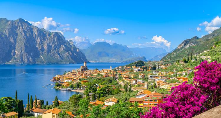 Vue classique de carte postale du Lac de Garde avec sa ville colorée, sa tour de château, les montagnes environnantes et les fleurs lumineuses au premier plan.