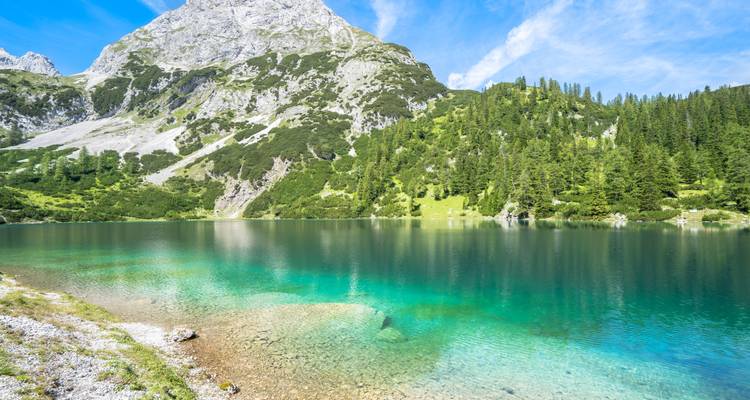 Lac alpin d'un turquoise cristallin reflétant la forêt de conifères et les versants rocheux des montagnes sous un ciel bleu éclatant.
