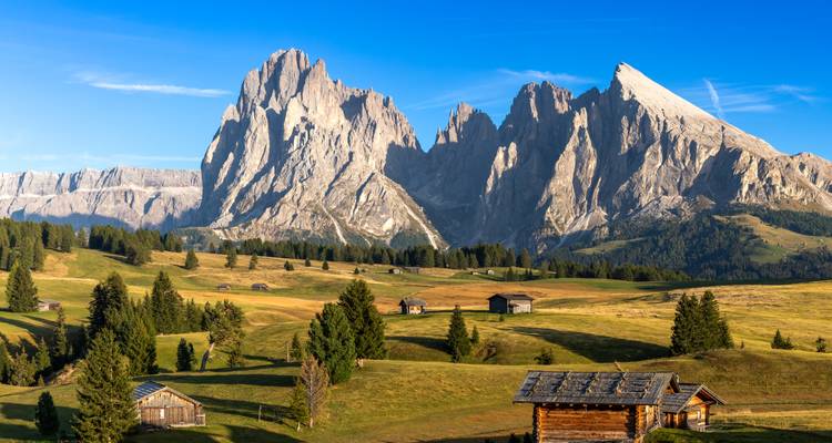 Les pics déchiquetés et dramatiques des Dolomites s'élevant au-dessus des prairies alpines ondulantes parsemées de chalets en bois à la lumière du matin.