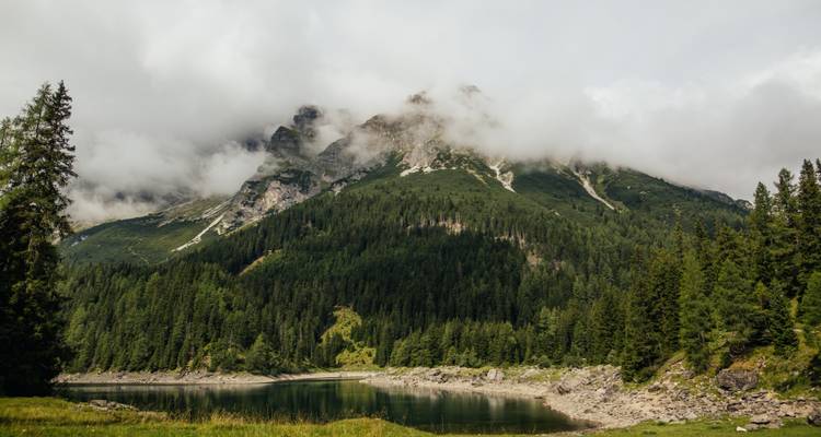 La niebla envuelve las laderas verdes de coníferas sobre un lago alpino reflectante bajo una cima parcialmente velada.