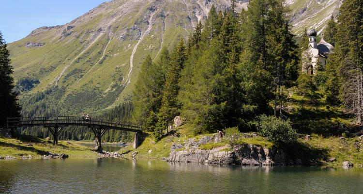 Un pequeño puente de madera cruza un tranquilo lago alpino con una capilla y empinadas laderas verdes de montaña al fondo.