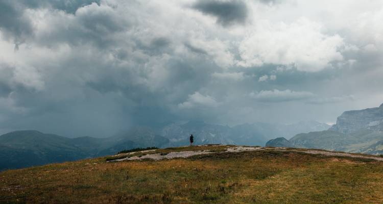 Una figura solitaria está de pie en una meseta alpina cubierta de hierba bajo unas nubes de tormenta espectaculares y montañas lejanas.