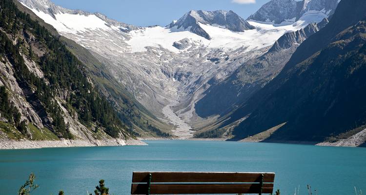 Bank mit Blick auf einen türkisfarbenen See und schneebedeckte Berge.