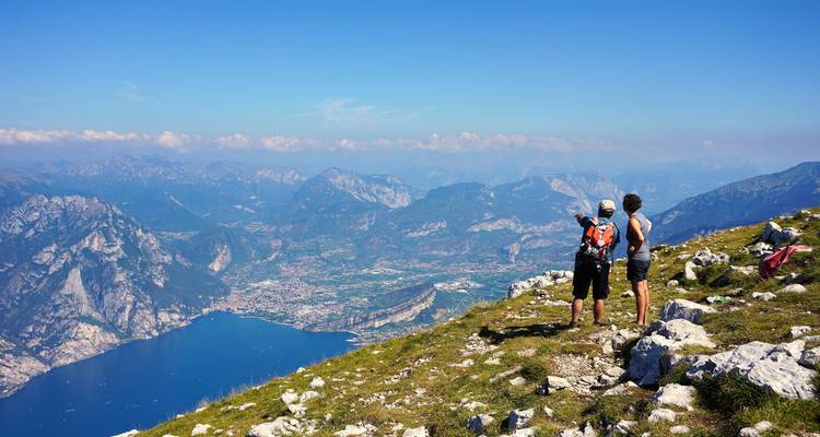 Zwei Wanderer stehen auf einem felsigen Gipfel mit Blick auf den Gardasee und entfernte Alpengipfel.