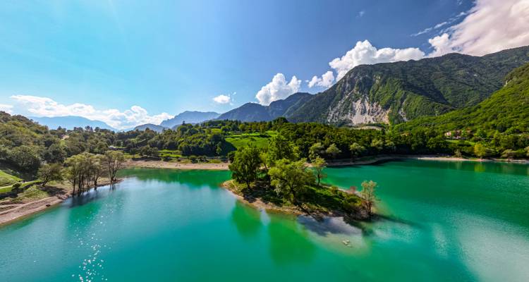 Smaragdgrüner Alpensee, umgeben von bewaldeten Hügeln und schneebedeckten Gipfeln unter einem klaren Himmel.