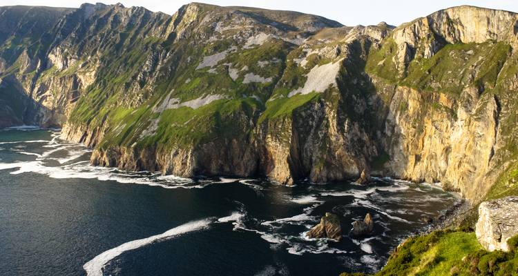 Sheer sea cliffs carpeted with green vegetation plunging into dark Atlantic waters with foamy surf.