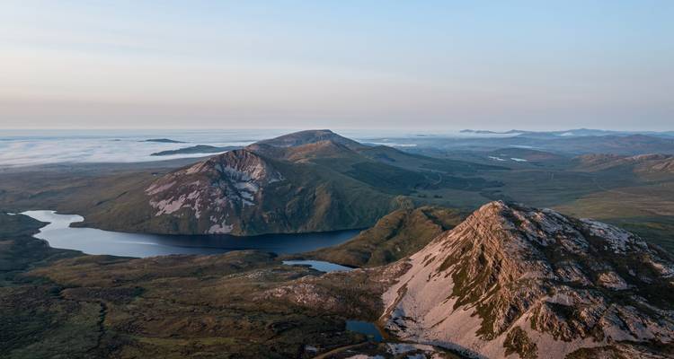 Aerial sunrise view over mist-filled valleys, lakes, and ridgelines of rugged Irish mountains.