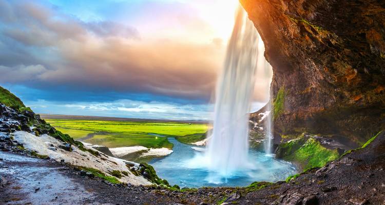 Ein mächtiger Wasserfall stürzt in ein türkisfarbenes Becken, das von üppigen grünen Ebenen und dramatischem Himmel eingefasst ist, der von warmen Sonnenstrahlen erleuchtet wird.