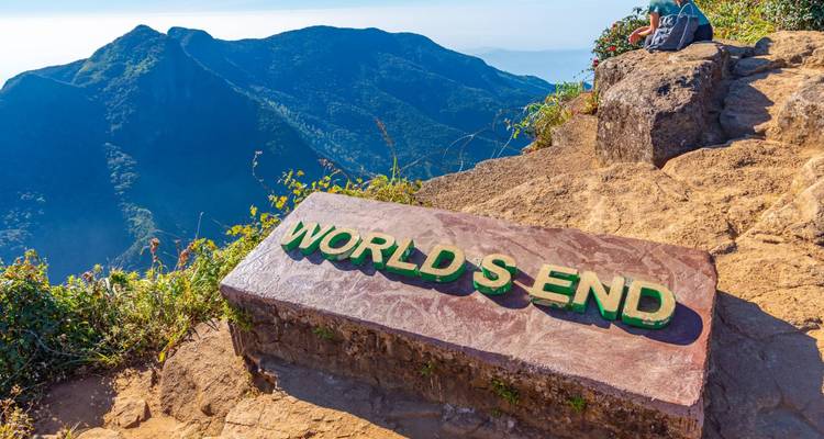 Marcador de piedra con letras verdes que dice 'WORLD'S END' ubicado en el borde de un acantilado con vista a montañas brumosas.