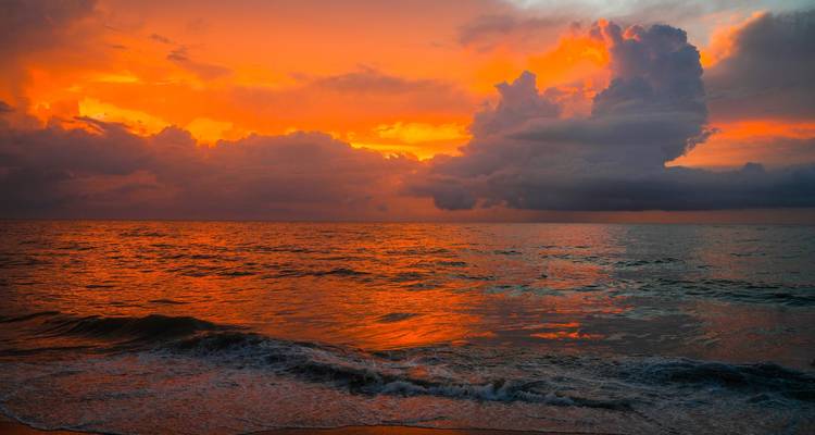 Puesta de sol naranja y roja vívida sobre el Océano Índico con nubes dramáticas y olas suaves.
