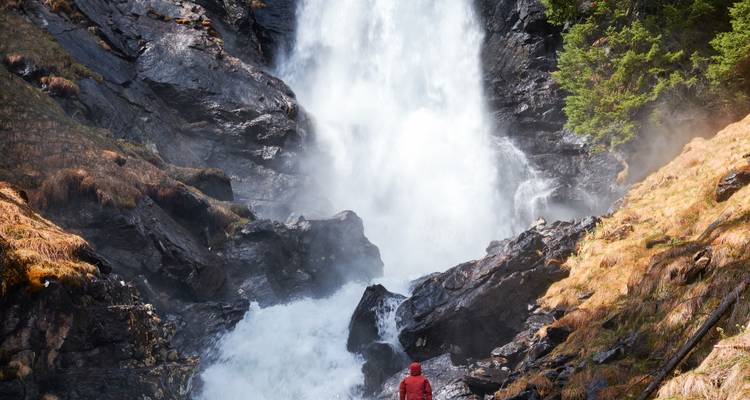 A figure in a red jacket facing a powerful waterfall cascading down a rocky cliff.