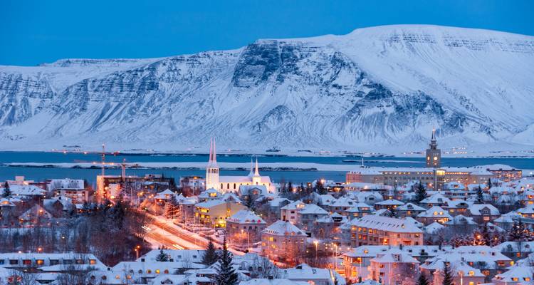Das schneebedeckte Reykjavik leuchtet unter dem Licht der Dämmerung in Blautönen, während der Berg Esja dahinter aufragt.