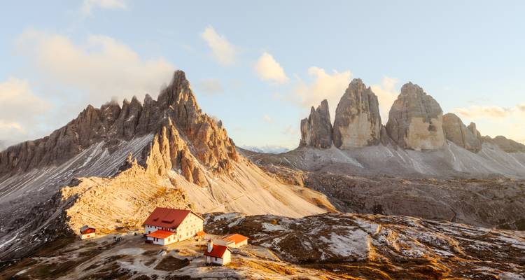 Ein Bergrefugium mit roten Dächern liegt unter sonnenbeleuchteten felsigen Gipfeln der Tre Cime im warmen Licht der goldenen Stunde.