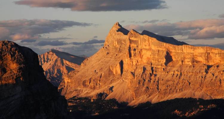 Goldensteindlicht badet einen dramatischen Dolomitengipfel und umgebende Klippen unter verstreuten Wolken.