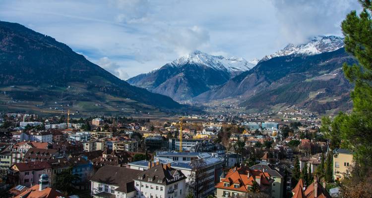 Panoramic view of Merano town surrounded by snow-capped Alps and green valleys below a partly cloudy sky.