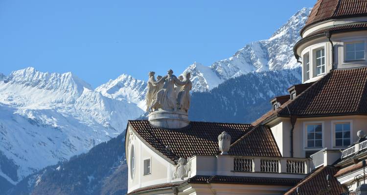 Detail of a stately building with classical sculptures and red-tiled roof set against snow-covered Alpine peaks under a clear blue sky.