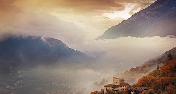 Castle surrounded by mist with autumn foliage and mountains.