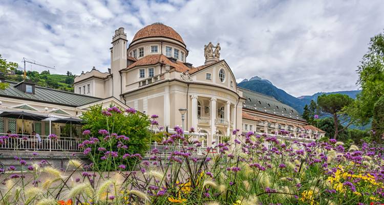 Elegante edificio histórico Kurhaus rodeado de vibrantes flores púrpuras y amarillas con las montañas alpinas de fondo.