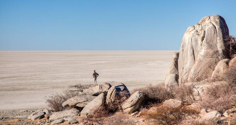 Voyageur solitaire debout sur des rochers surplombant la vaste étendue blanche des salines du Makgadikgadi sous un ciel sans nuages.