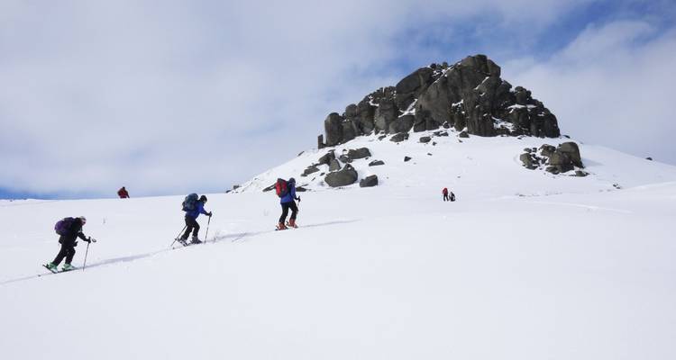 Skifahrer, die einen verschneiten Hang zu einem felsigen Gipfel hinaufsteigen.