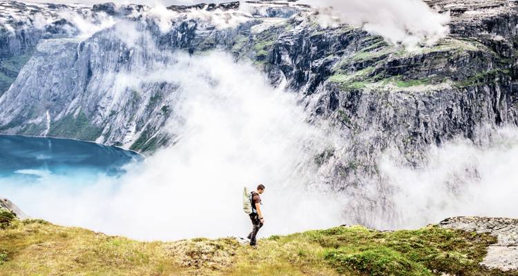 Wanderer, der auf einem grasbewachsenen Bergrücken steht und auf einen Fjord und Berge blickt.