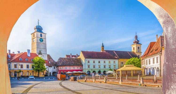 Place de ville animée encadrée par une arche, avec des bâtiments historiques et une tour d'horloge.