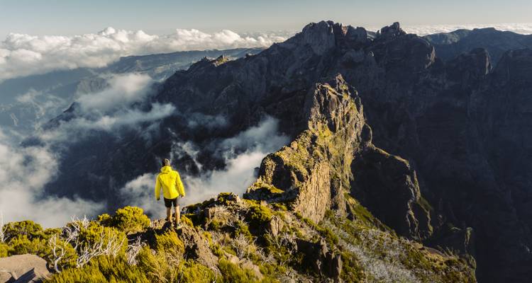 Een eenzame wandelaar in een geel jack staat op een dramatische, door wolken omgeven bergrug in de hooglanden van Madeira.