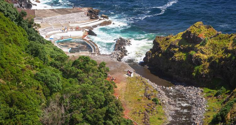 Uitzicht vanaf de kliffetop op een natuurlijk zwembadcomplex gevoed door de oceaan, verscholen tussen rotsachtige oevers en groene hellingen.
