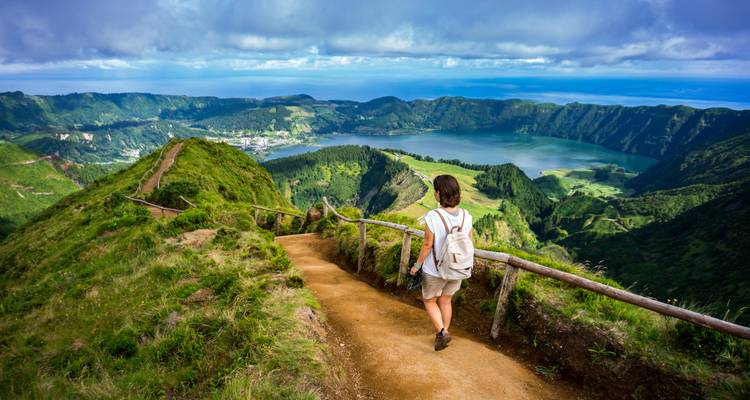 Een jonge wandelaar met een rugzak loopt over een zandpad met uitzicht op de twee kraatermeren van Sete Cidades, omgeven door weelderige groene caldeirawanden en de Atlantische Oceaan in de verte.