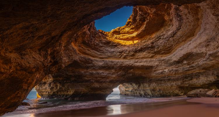 Sunlit interior of the Benagil sea cave with golden walls opening to blue sky and ocean.