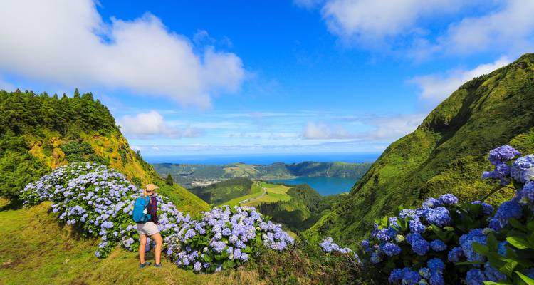 Wandelaar staat naast bloeiende blauwe hortensia's met uitzicht op de kratermeren van Sete Cidades en de heuvels.