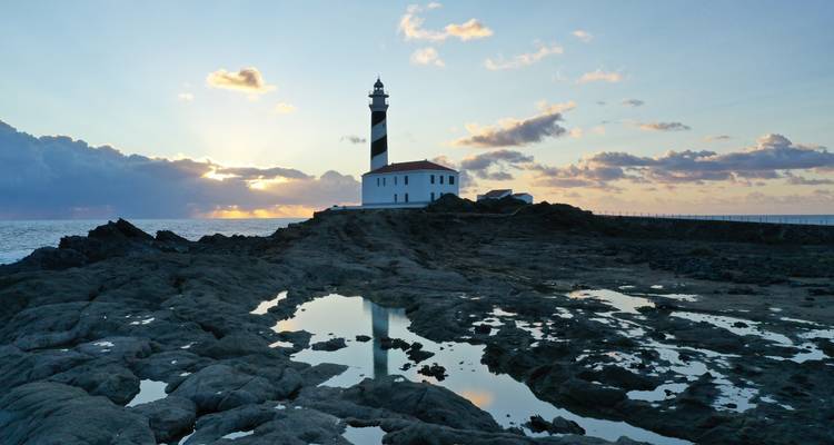 Faro en costa rocosa al atardecer con reflejos en pozas de marea.