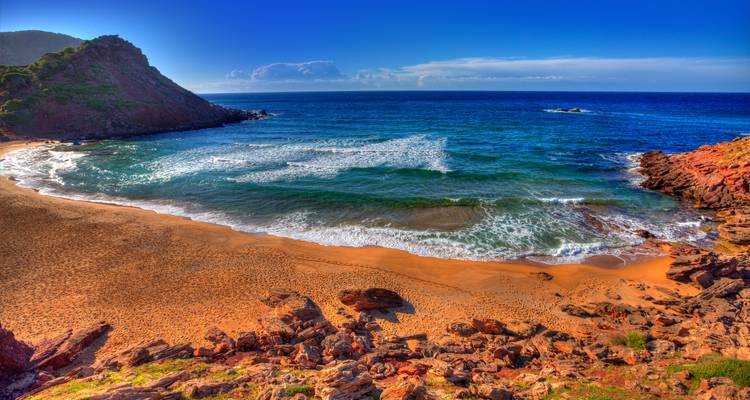 Playa rocosa con agua azul clara y olas rompiendo en la orilla.