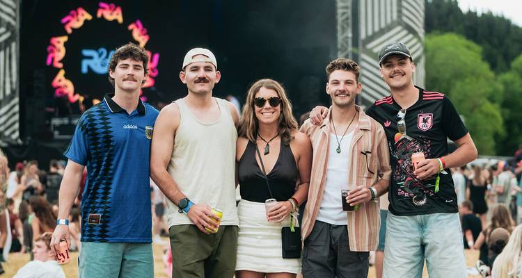 Five friends pose at an outdoor music festival stage holding drinks amid a lively crowd.