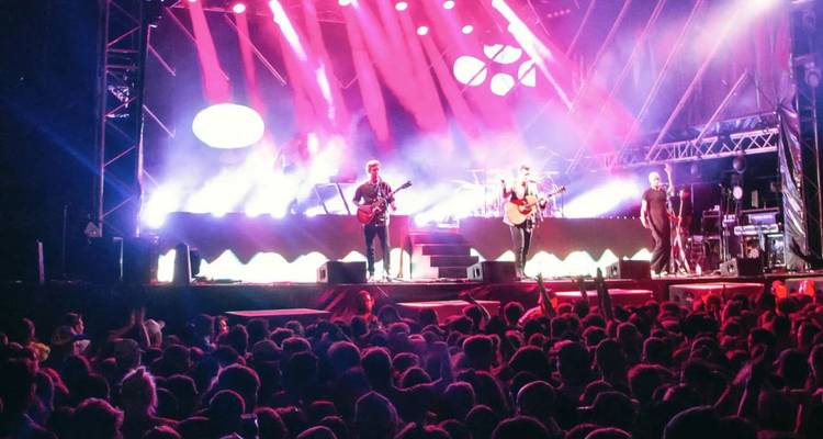 Scène de festival de musique en plein air avec des lumières roses vives illuminant une grande foule qui acclame lors d'une soirée.