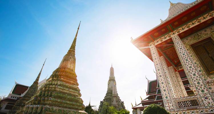 Les flèches et stupas illuminés par le soleil d'un complexe de temples à Bangkok, photographiés sous un angle bas contre un ciel bleu dégagé.